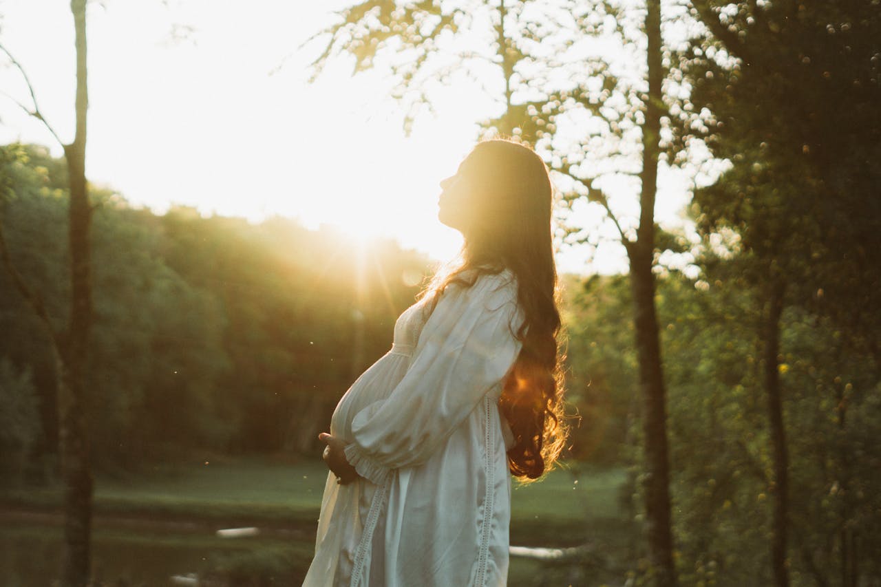 discover Pregnant woman in white dress enjoying a sunset in a serene forest setting.