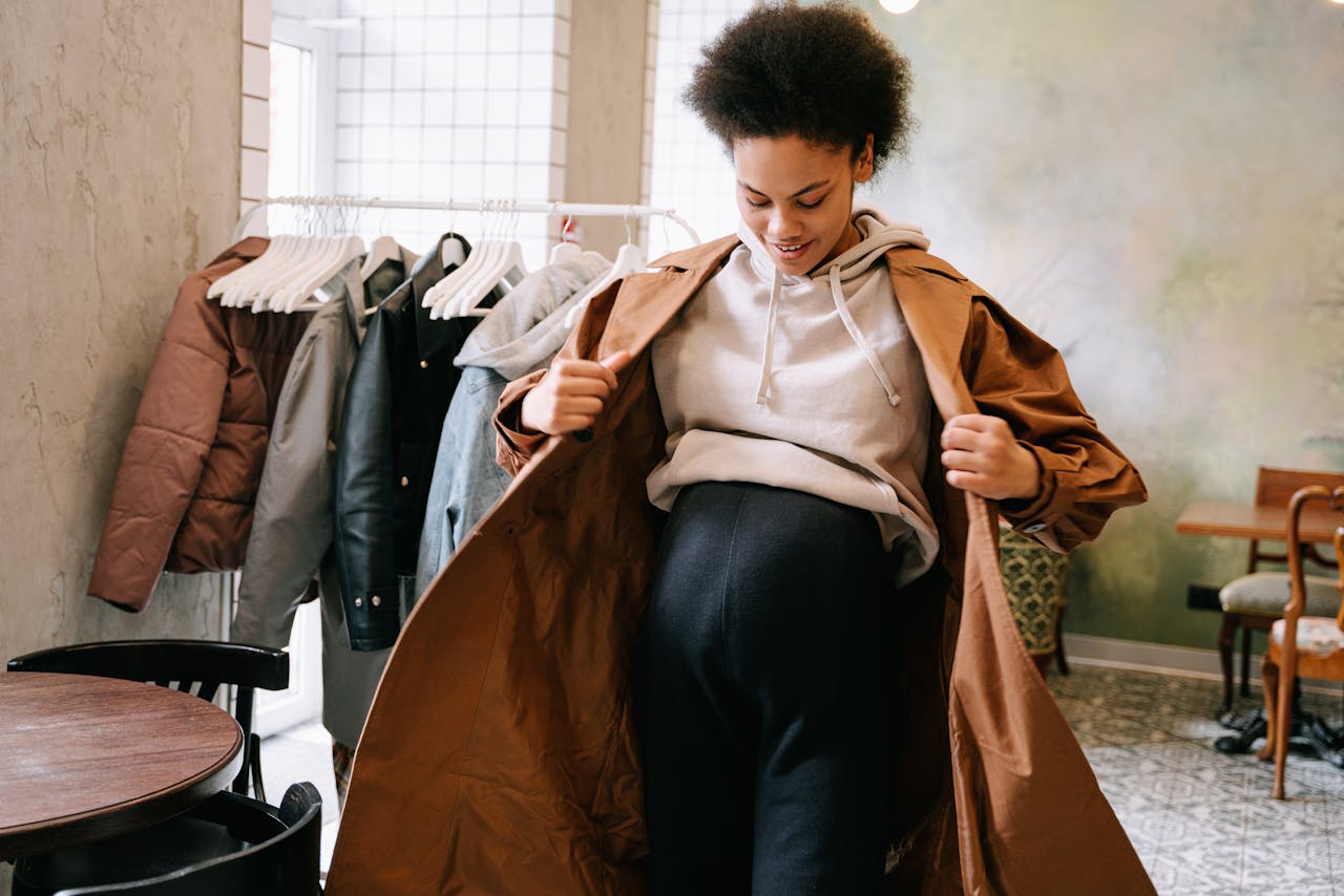 about-us Pregnant woman in stylish coat adjusting outfit indoors near clothes rack.