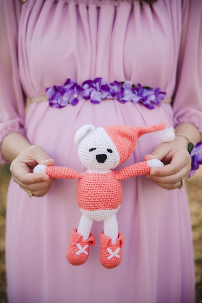 Pregnant woman in lavender dress holding cute knitted teddy bear, symbolizing motherhood and anticipation.
